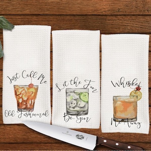 May include: Three white kitchen towels with cocktail illustrations and text. The first says "Just Call Me Old Fashioned", the second "Let the Fun Be-Gin", and the third "Whiskey Me Away". A stainless steel knife with a wooden handle is in the foreground.