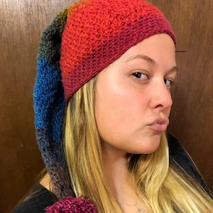 Puede incluir: Una mujer lleva un gorro de crochet de colores con una cola larga y puntiaguda y un pompón en el extremo. El gorro está hecho de diferentes tonos de rojo, naranja, amarillo, verde, azul y morado.
