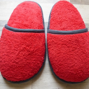 Puede incluir: Un par de zapatillas de casa de color rojo brillante confeccionadas en un tejido de rizo suave y texturizado. Cada zapatilla presenta una puntera redondeada y un ribete gris oscuro que perfila la abertura y la suela, creando un contraste.