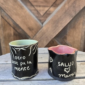 May include: Two handmade ceramic shot glasses. One is black with white text that reads "otro shot pa la mente" and a green interior. The other is black with white text that reads "SALUD Mami" and a pink interior.