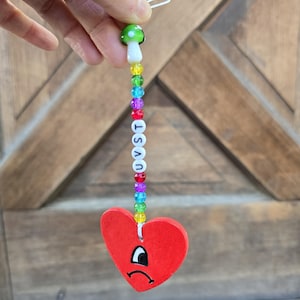 May include: A handmade charm featuring a red heart with a sad face, strung with colorful beads and the letters UVST. The charm is topped with a green and white mushroom bead and a white loop.