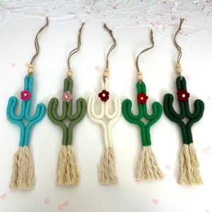 May include: Five macrame cactus ornaments in varying colors: turquoise, olive green, cream, green, and dark green. Each cactus features a small flower and a tassel. They are suspended by twine and wooden beads, perfect for adding a touch of desert charm.