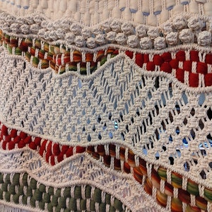 May include: A white macrame wall hanging with red, green, and orange woven details. The hanging has a fringe at the bottom.