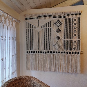 Puede incluir: Un gran tapiz de macramé en crema y negro con diseño geométrico. La decoración de pared presenta patrones de diamantes, rayas y flecos. La pieza está montada en una barra de madera y cuelga de una pared blanca.
