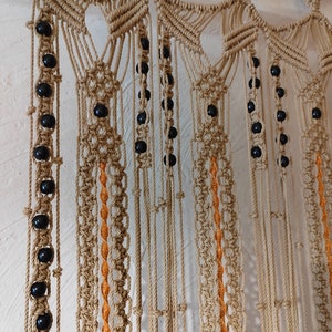 Peut inclure: Une suspension murale en macramé avec un motif géométrique et des perles noires. La suspension est réalisée avec un cordon de couleur naturelle et possède un  tringle en bois brun clair en haut.
