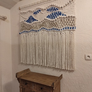 Peut inclure: Une tapisserie murale en macramé blanche avec des accents bleus et beiges. La tapisserie présente un motif de vagues et des franges. Elle est exposée sur un mur blanc au-dessus d'une armoire en bois.
