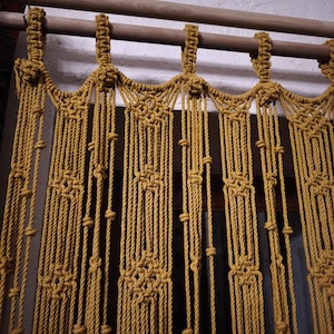 Peut inclure: Un rideau en macramé jaune moutarde avec une tringle en bois en haut. Le rideau est fait d'une corde épaisse et présente un motif géométrique.