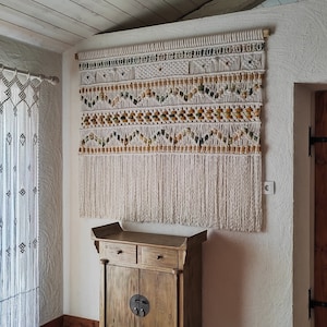 Peut inclure: Une tapisserie murale en macramé blanche avec un motif géométrique et des perles brunes. La tapisserie est fixée sur un mur blanc au-dessus d'une armoire en bois avec deux tiroirs et deux portes.