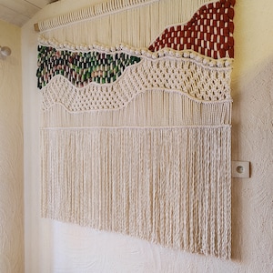 Peut inclure: Une grande tapisserie murale en macramé avec un motif tissé présentant une combinaison de fils de couleur crème naturelle, verte et rouge. La tapisserie a une frange de fils de couleur crème naturelle.