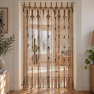 Puede incluir: Una cortina de puerta de macramé hecha de cuerda beige natural, con intrincados patrones de nudos, diseños en forma de diamante y cuentas de color verde oscuro y rojo intercaladas. La cortina cuelga de una barra y termina en flecos.
