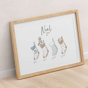 Op de afbeelding: Een witte ingelijste print met het woord "Noël" en het jaar "2021" in zwarte schrijfletters. Vier blauwe en witte kousen hangen met namen onder elke kous: Julien, Marc, Valentin en Camille.