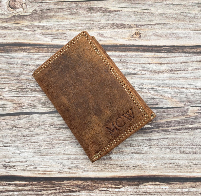 May include: A brown leather wallet with visible stitching and the initials "MCW" embossed on the front. The wallet has a worn, distressed appearance, suggesting a vintage or rustic style. The leather is a warm, earthy brown colour.
