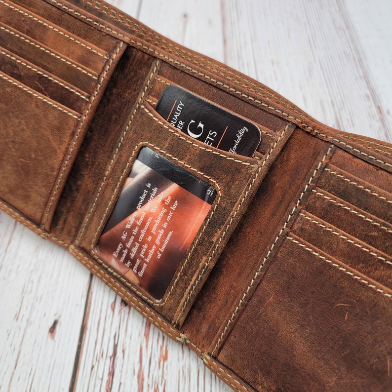 May include: Open brown leather wallet with multiple card slots and a bill compartment. The wallet has white stitching and contains two cards. One card reads "QUALITY LEATHER GOODS" and the other card has text that is partially visible.