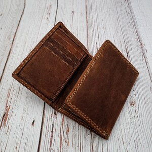 May include: Open brown leather wallet with multiple card slots and a bill compartment. The wallet is made of distressed leather with visible stitching around the edges. The wallet is open, displaying the interior and the card slots.