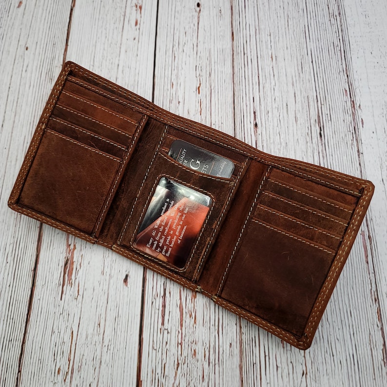 May include: Open brown leather wallet with several card slots and a clear ID window. The wallet is made of distressed leather and has white stitching around the edges. A card with text is visible inside the wallet.