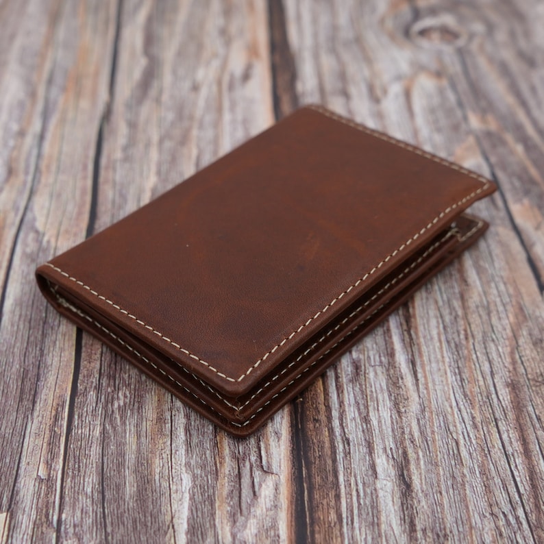 May include: A brown leather wallet with white stitching. The wallet is folded closed and is lying on a wooden surface.