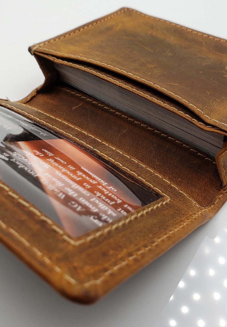 May include: A brown leather card holder with a stitched edge and a clear plastic window for displaying a business card. The card holder is open and shows the interior with multiple card slots.