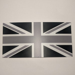 Puede incluir: Una pegatina de bandera Union Jack en blanco y negro. La bandera está impresa sobre un fondo blanco.