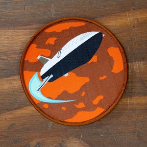 Könnte beinhalten: Runder, gestickter Aufnäher mit einem weißen und schwarzen Space Shuttle-Design vor einem orange-braunen Hintergrund. Eine hellblaue Spur ist sichtbar. Der Aufnäher hat einen braunen Rand.