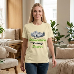 May include: A cream-coloured short-sleeve t-shirt featuring a graphic of a mountain range with pine trees and a silhouette of a person and dog. The text reads The Mountains Are Calling Jasper in a mix of green and black script and block lettering.