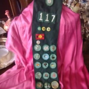 May include: A green sash with a number "117" and various badges and patches. The sash is displayed on a pink fabric background.