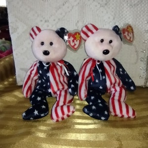 May include: Two plush beanie baby bears wearing red, white, and blue patriotic outfits. The bears have pink noses and black eyes. The bears are sitting on a gold surface.