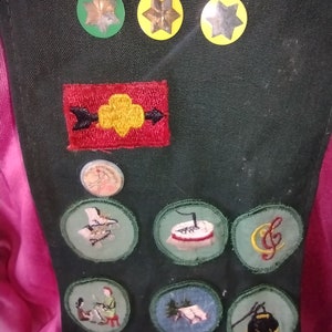 May include: A green Girl Scout sash with various embroidered patches, including a yellow trefoil, a red arrow, and a music note. There are also three yellow stars with gold centers.