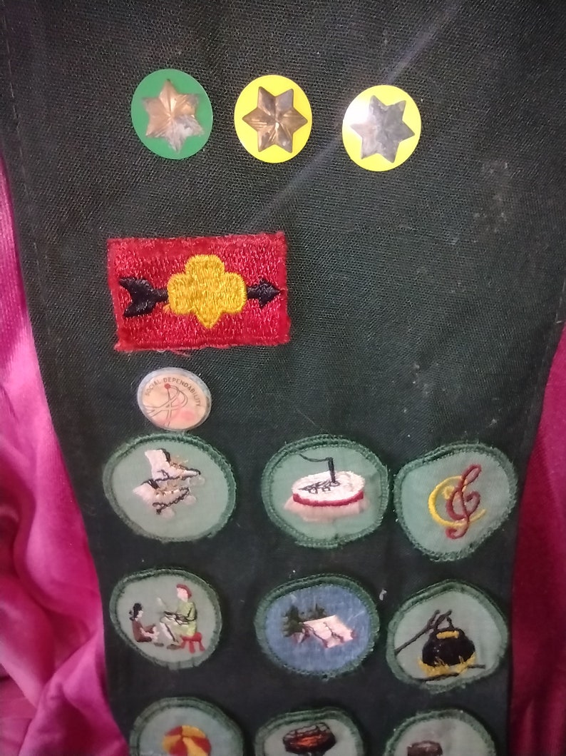 May include: A green Girl Scout sash with various embroidered patches, including a yellow trefoil, a red arrow, and a music note. There are also three yellow stars with gold centers.