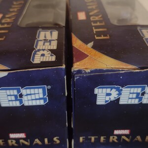 Puede incluir: Dos cajas de dispensadores PEZ con el logo de Marvel Eternals. Las cajas son de color azul oscuro con letras doradas y blancas. El logotipo de PEZ se muestra en blanco y azul.
