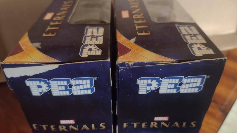 Puede incluir: Dos cajas de dispensadores PEZ con el logo de Marvel Eternals. Las cajas son de color azul oscuro con letras doradas y blancas. El logotipo de PEZ se muestra en blanco y azul.