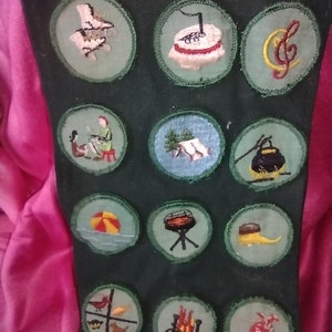 May include: A green fabric sash with various embroidered patches. The patches feature images of a campfire, a musical note, a tent, a beach ball, a grill, a bird, a dog, a pot, a shoe, a bag, and a girl playing a piano.