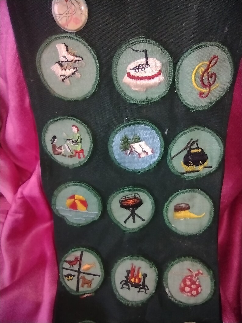 May include: A green fabric sash with various embroidered patches. The patches feature images of a campfire, a musical note, a tent, a beach ball, a grill, a bird, a dog, a pot, a shoe, a bag, and a girl playing a piano.