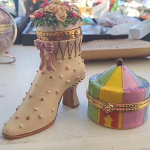 May include: A decorative ceramic boot-shaped trinket box with a floral arrangement on top. The boot is cream-coloured with pink and gold accents. A small, colourful circus-tent-shaped trinket box sits beside it.
