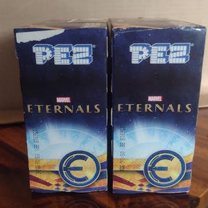 Puede incluir: Dos cajas de dispensadores de caramelos PEZ con el logotipo de Marvel Eternals. Las cajas son de color azul oscuro con un fondo estrellado y un dise&ntilde;o circular dorado y azul. El logotipo de PEZ est&aacute; en la parte superior.