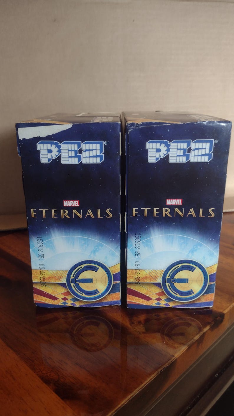 Puede incluir: Dos cajas de dispensadores de caramelos PEZ con el logotipo de Marvel Eternals. Las cajas son de color azul oscuro con un fondo estrellado y un dise&ntilde;o circular dorado y azul. El logotipo de PEZ est&aacute; en la parte superior.