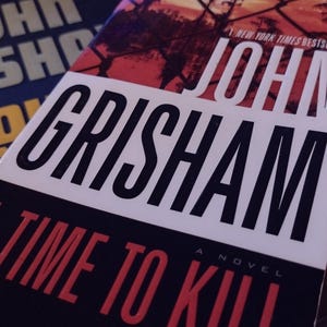 May include: A paperback book titled "A Time To Kill" by John Grisham. The cover features a red and white background with a black fence and a black and white text overlay.