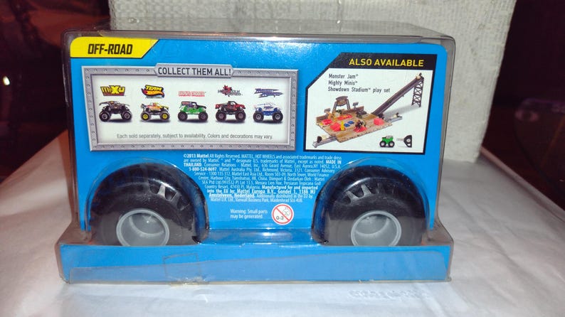May include: A blue and white cardboard box containing a Monster Jam truck toy. The box features an image of the truck on the front, along with the text "Off-Road" and "Collect Them All!" There's also a picture of a Monster Jam playset with the words "Monster Jam Mighty Minis Showdown Stadium play set".