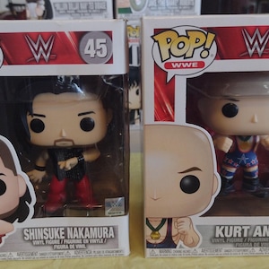 Puede incluir: Dos figuras de vinilo Funko Pop! WWE en sus cajas originales. Una es Shinsuke Nakamura, la otra es Kurt Angle. Las cajas son blancas con detalles rojos y presentan el logotipo de la WWE y los nombres de los personajes.