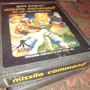 Puede incluir: Un cartucho de juego Atari negro con una etiqueta colorida que presenta una ilustración de dibujos animados de un piloto en una cabina. La etiqueta dice "game program missile command" y "Use with Joystick Controllers". El cartucho también tiene una etiqueta negra en el lateral que dice "missile command".