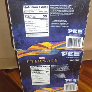 Puede incluir: Dos cajas de caramelos PEZ apiladas con el logotipo de Marvel Eternals. Las cajas tienen un esquema de color azul y dorado con informaci&oacute;n nutricional e ingredientes. El logotipo de PEZ y la direcci&oacute;n del sitio web son visibles.