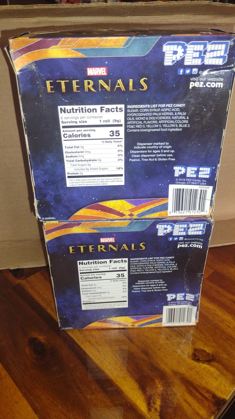 Puede incluir: Dos cajas de caramelos PEZ apiladas con el logotipo de Marvel Eternals. Las cajas tienen un esquema de color azul y dorado con informaci&oacute;n nutricional e ingredientes. El logotipo de PEZ y la direcci&oacute;n del sitio web son visibles.