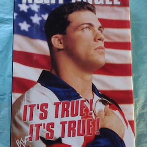 Puede incluir: Portada de libro con Kurt Angle frente a la bandera estadounidense. El texto "KURT ANGLE" está en la parte superior, y "IT'S TRUE! IT'S TRUE!" se muestra en rojo. El logotipo de la WWE es visible.