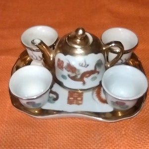 Op de afbeelding: Een wit porseleinen theeservies met gouden accenten. De theepot heeft een drakenontwerp en de woorden "Dubbel geluk" in Chinese karakters. De set bevat vier kleine kopjes en een dienblad.