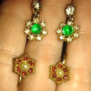 Pode incluir: Brincos dourados com uma pedra verde central rodeada por cristais transparentes. Um segundo par de brincos com um design de flor de pedra vermelha e um centro de pérola também é visível.