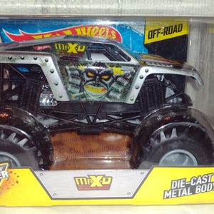 May include: A silver and black Monster Jam die-cast toy truck with the name "Max-D" on the side. The truck is in a clear plastic package with the words "Hot Wheels", "Off-Road", "Monster Jam", and "Die-Cast Metal Body" on the packaging. The truck is suitable for ages 3 and up.