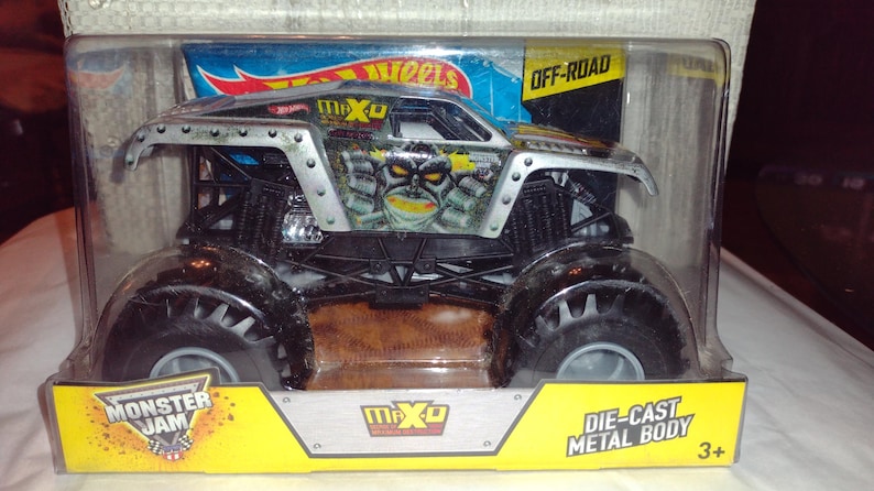 May include: A silver and black Monster Jam die-cast toy truck with the name "Max-D" on the side. The truck is in a clear plastic package with the words "Hot Wheels", "Off-Road", "Monster Jam", and "Die-Cast Metal Body" on the packaging. The truck is suitable for ages 3 and up.