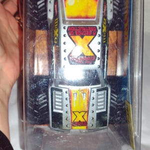 May include: A silver and black monster truck toy with a yellow and red flame design on the hood. The truck is in a clear plastic package and has the text "2 GENERATIONS OF DESTRUCTION" on the side.