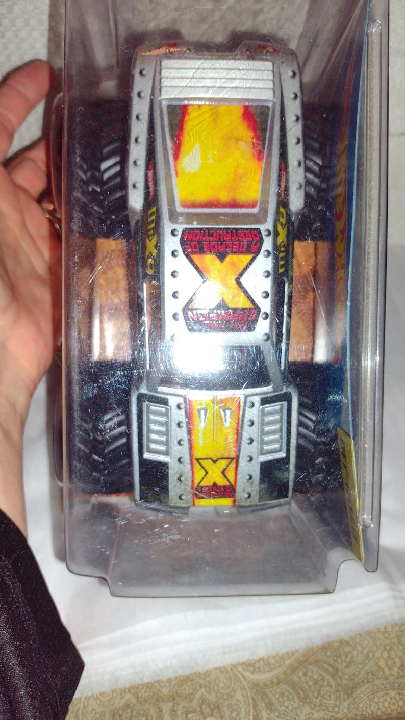 May include: A silver and black monster truck toy with a yellow and red flame design on the hood. The truck is in a clear plastic package and has the text "2 GENERATIONS OF DESTRUCTION" on the side.
