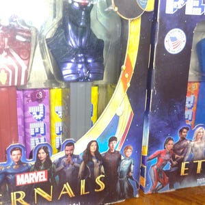 Puede incluir: Dispensadores de PEZ de Marvel Eternals en embalaje. Los dispensadores presentan cabezas de personajes en azul, verde y morado. El embalaje tiene un fondo azul oscuro con im&aacute;genes de personajes y el logotipo de Marvel Eternals. Fabricado en EE. UU.