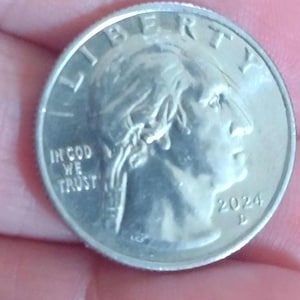 May include: A silver quarter coin with the profile of a person facing right. The coin has the words "LIBERTY", "IN GOD WE TRUST", and the year "2024" visible. The coin is held in a hand.